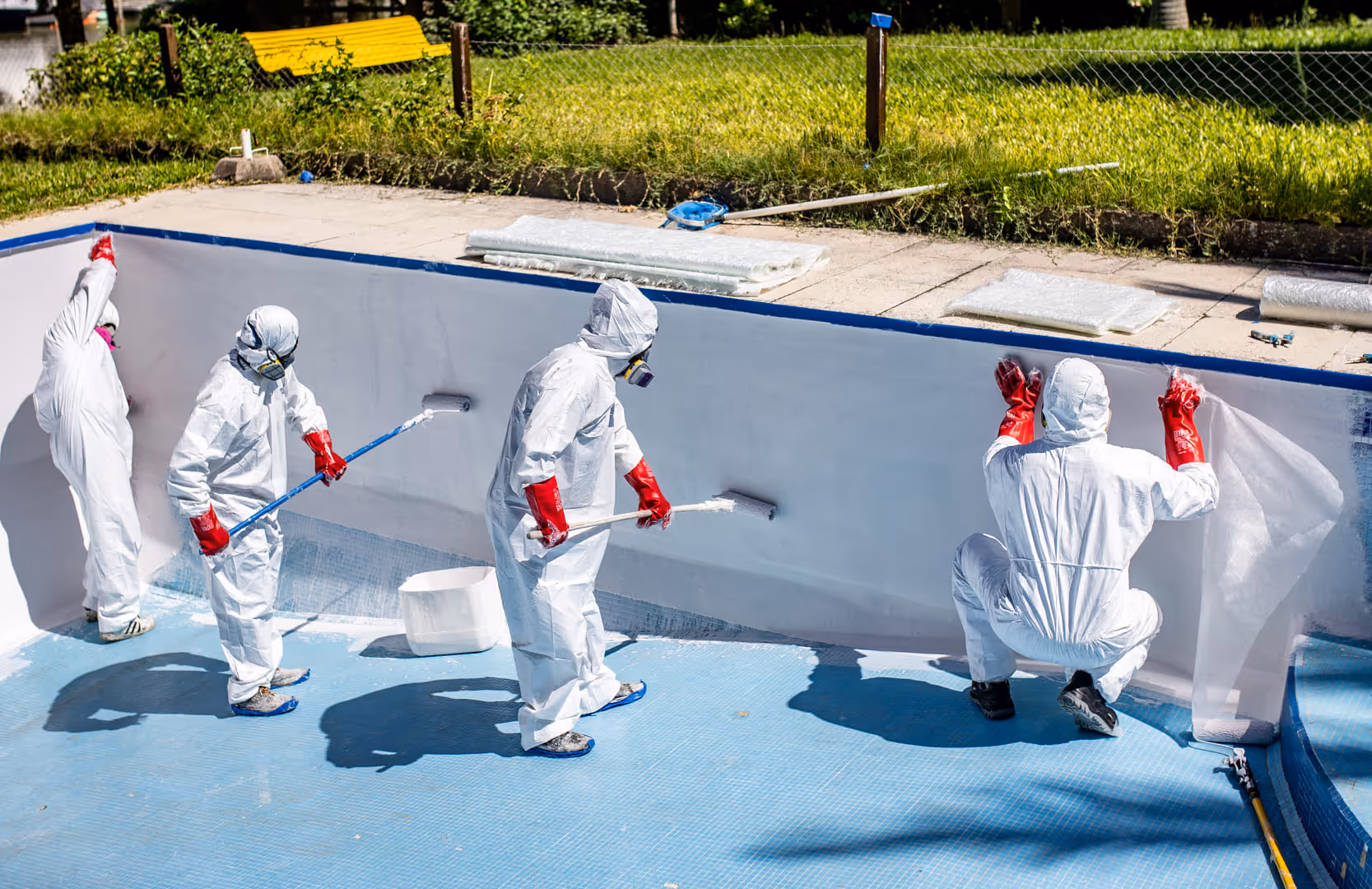 Cuatro trabajadores con trajes protectores blancos y guantes rojos aplican una capa blanca dentro de una piscina vacía.