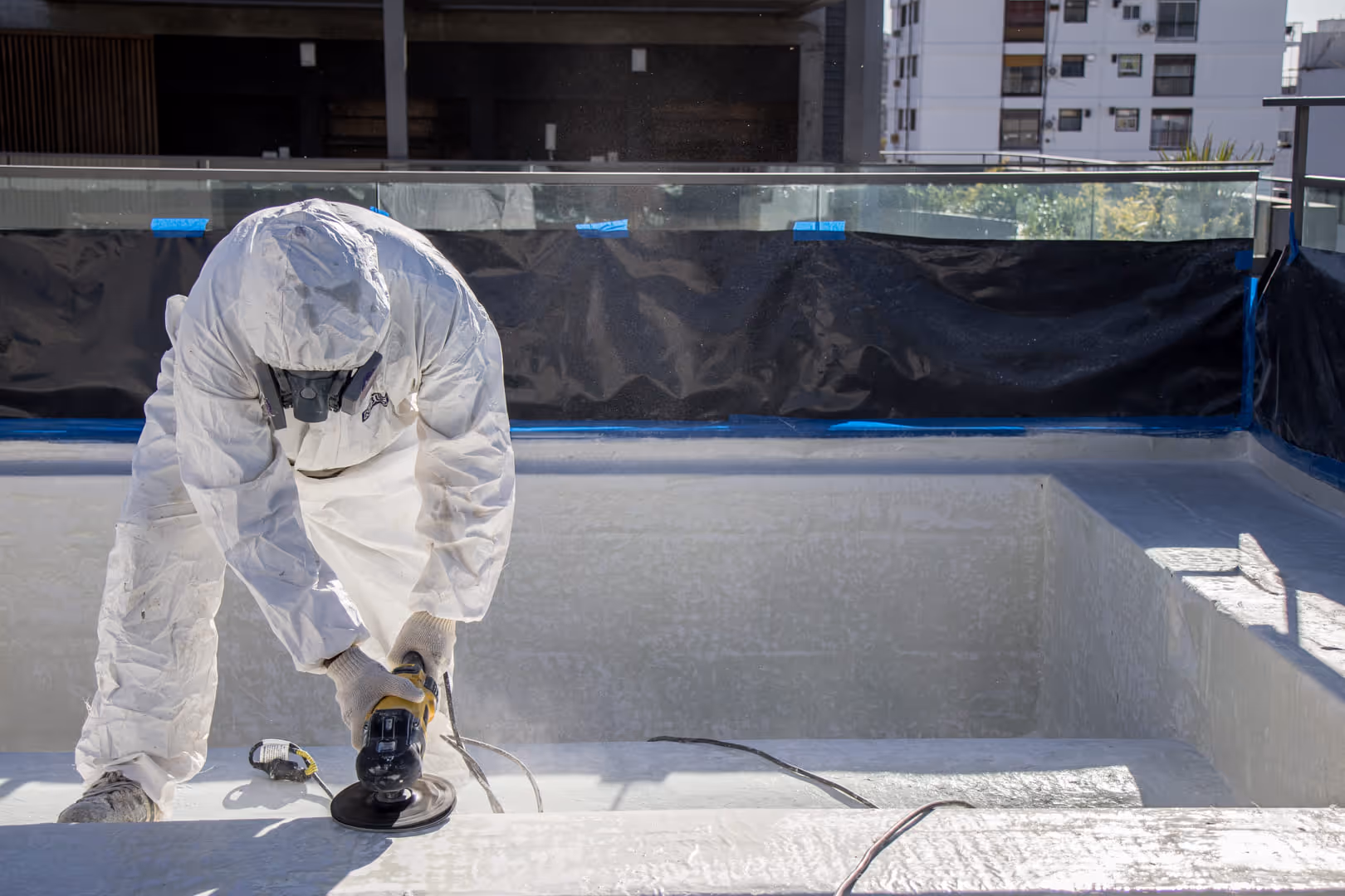 Trabajador con traje protector blanco y máscara utilizando una herramienta eléctrica para pulir o esmerilar la superficie interior de una piscina vacía.