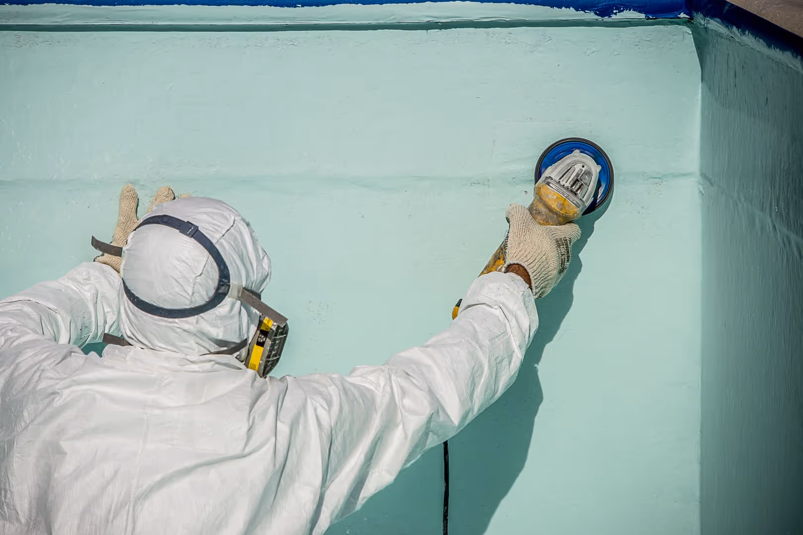 Persona con traje de protección y guantes utilizando una herramienta eléctrica para lijar una pared de color verde claro.