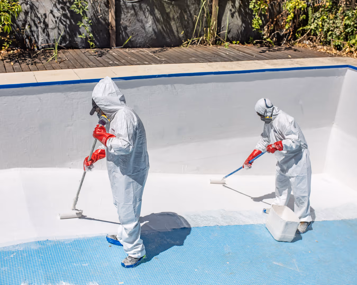 Dos trabajadores con trajes protectores blancos, guantes rojos y máscaras pintan de blanco el interior de una piscina vacía con rodillos de pintura.