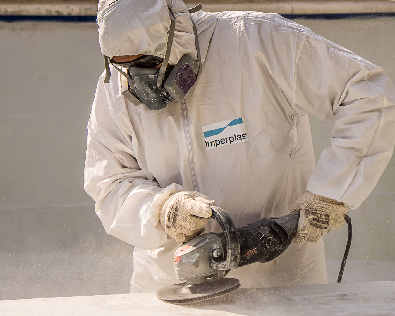 Trabajador con traje de protección y guantes utilizando una amoladora eléctrica sobre una superficie de hormigón, usando una máscara respiratoria.