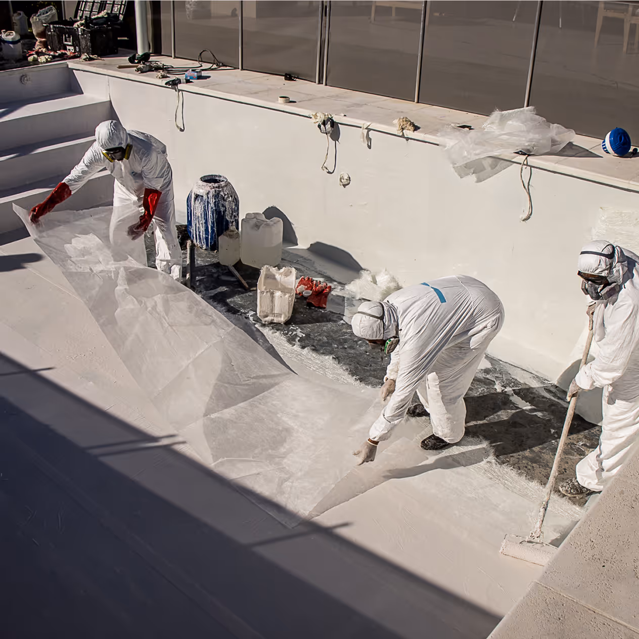Tres trabajadores con trajes de protección y guantes rojos aplican material de fibra de vidrio dentro de una piscina vacía.