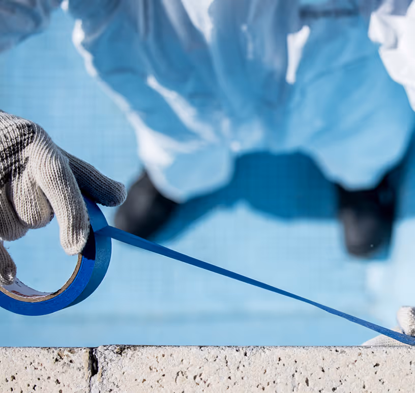 Persona con traje de protección y guantes aplicando cinta de pintor azul en el borde de una superficie cerca de una piscina.