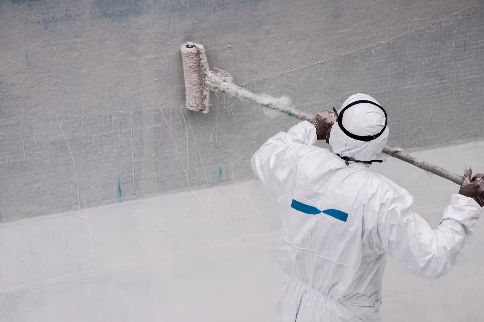 Trabajador con traje protector blanco y máscara puliendo o lijando la superficie interior de una piscina vacía con una herramienta eléctrica.