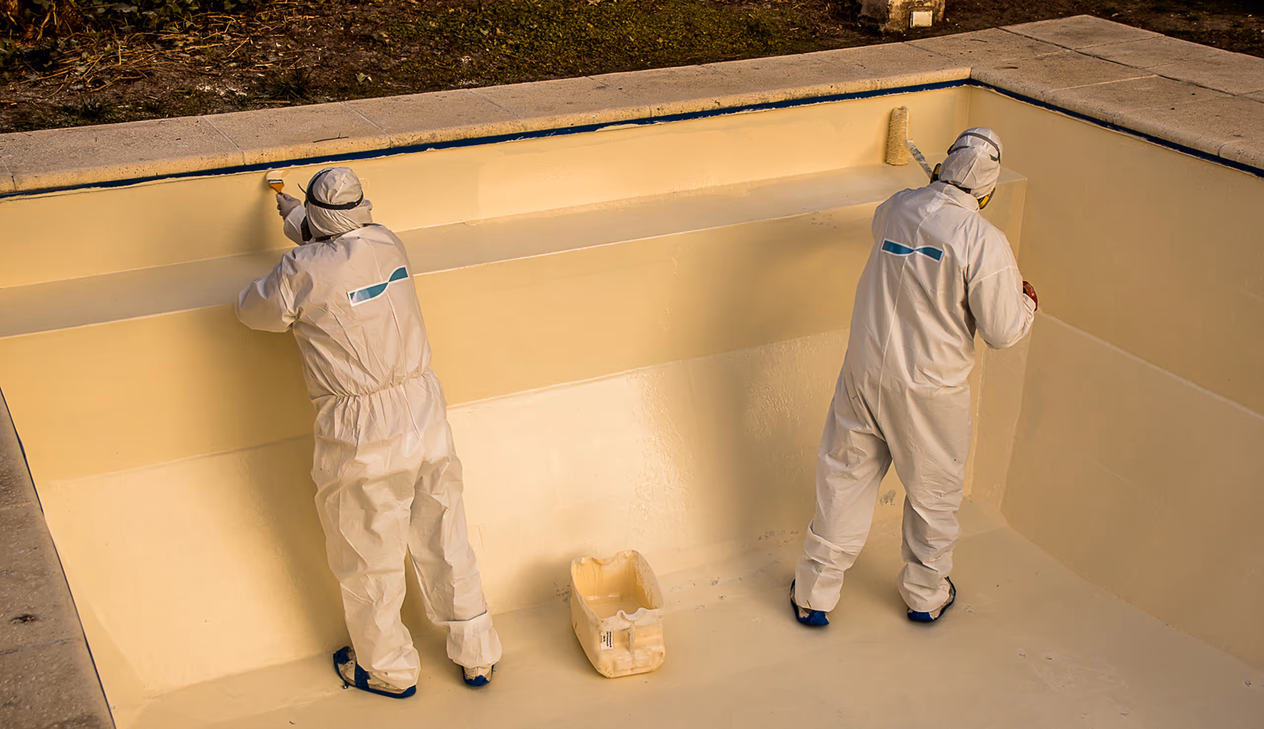 Dos trabajadores con trajes protectores blancos pintan la pared interior de una piscina vacía con pintura de color amarillo claro.