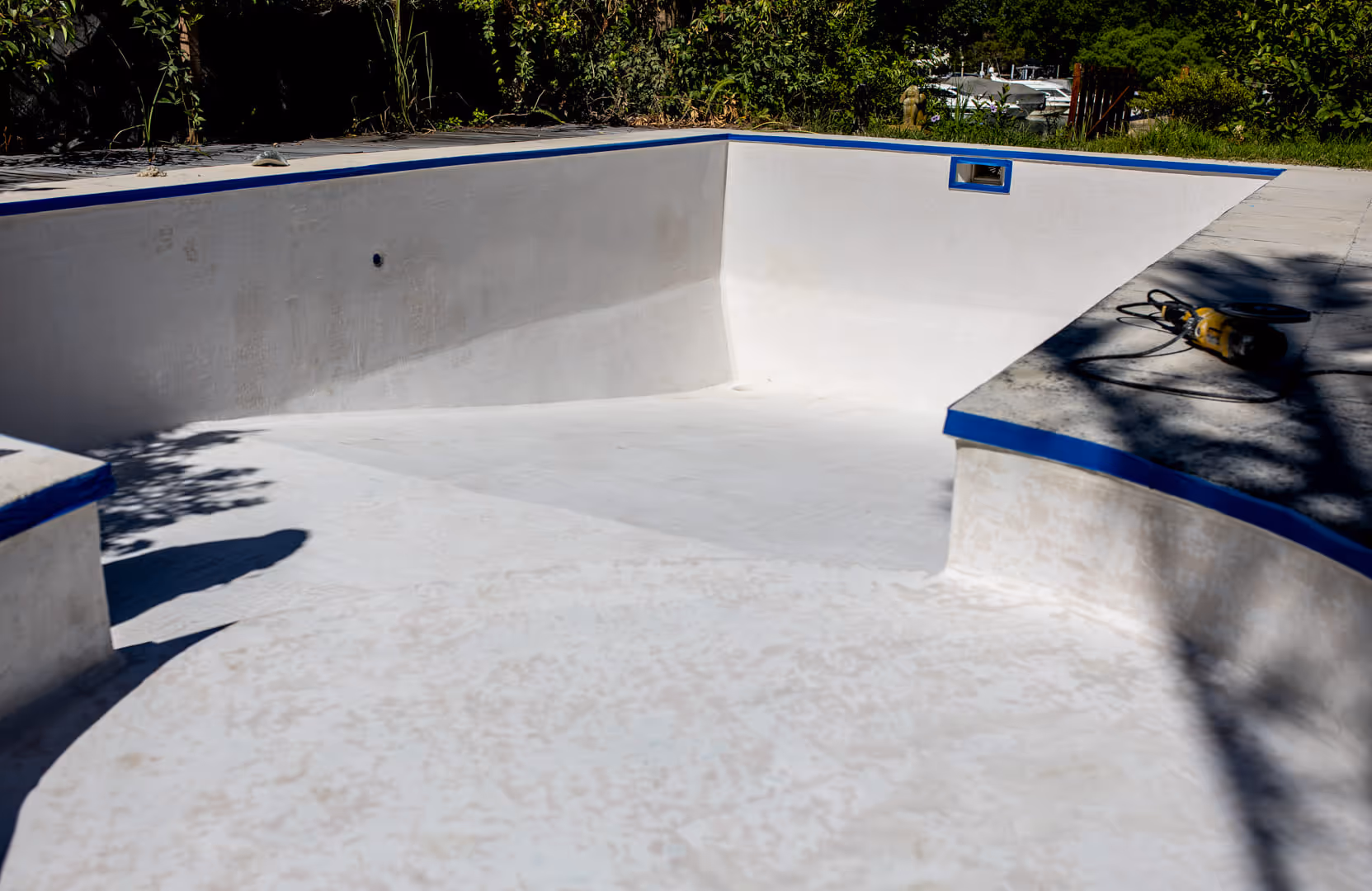 Piscina de hormigón blanco vacía en construcción con borde azul y una herramienta eléctrica en el borde.