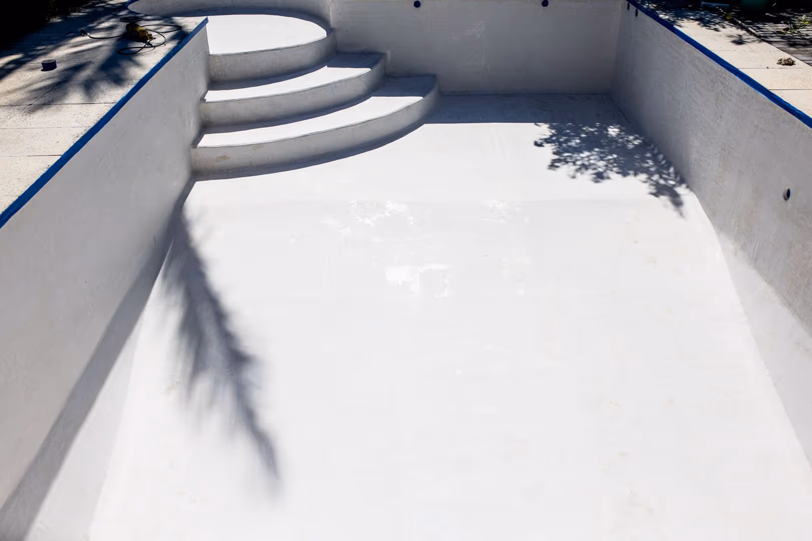 Piscina blanca vacía con escalones curvos y detalles azules bajo la brillante luz del sol.