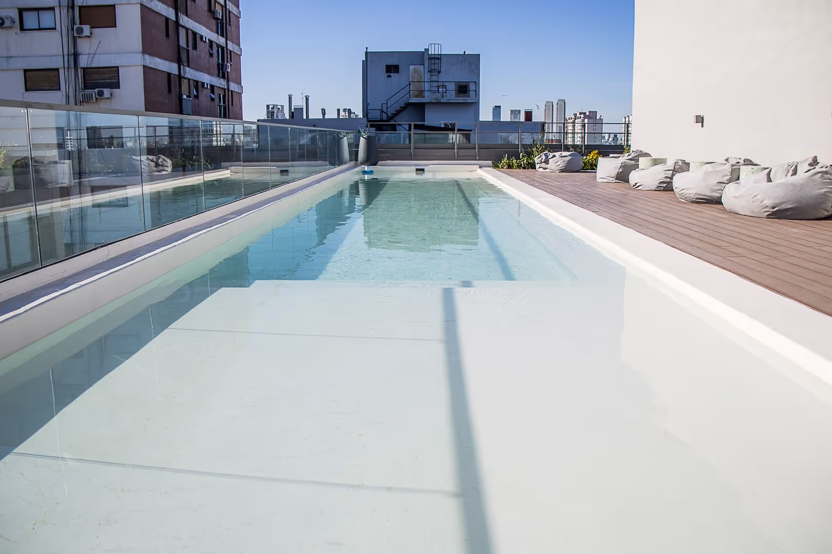 Piscina en la azotea con agua cristalina, barandilla de cristal a la izquierda y sillones puff grises sobre terraza de madera a la derecha.