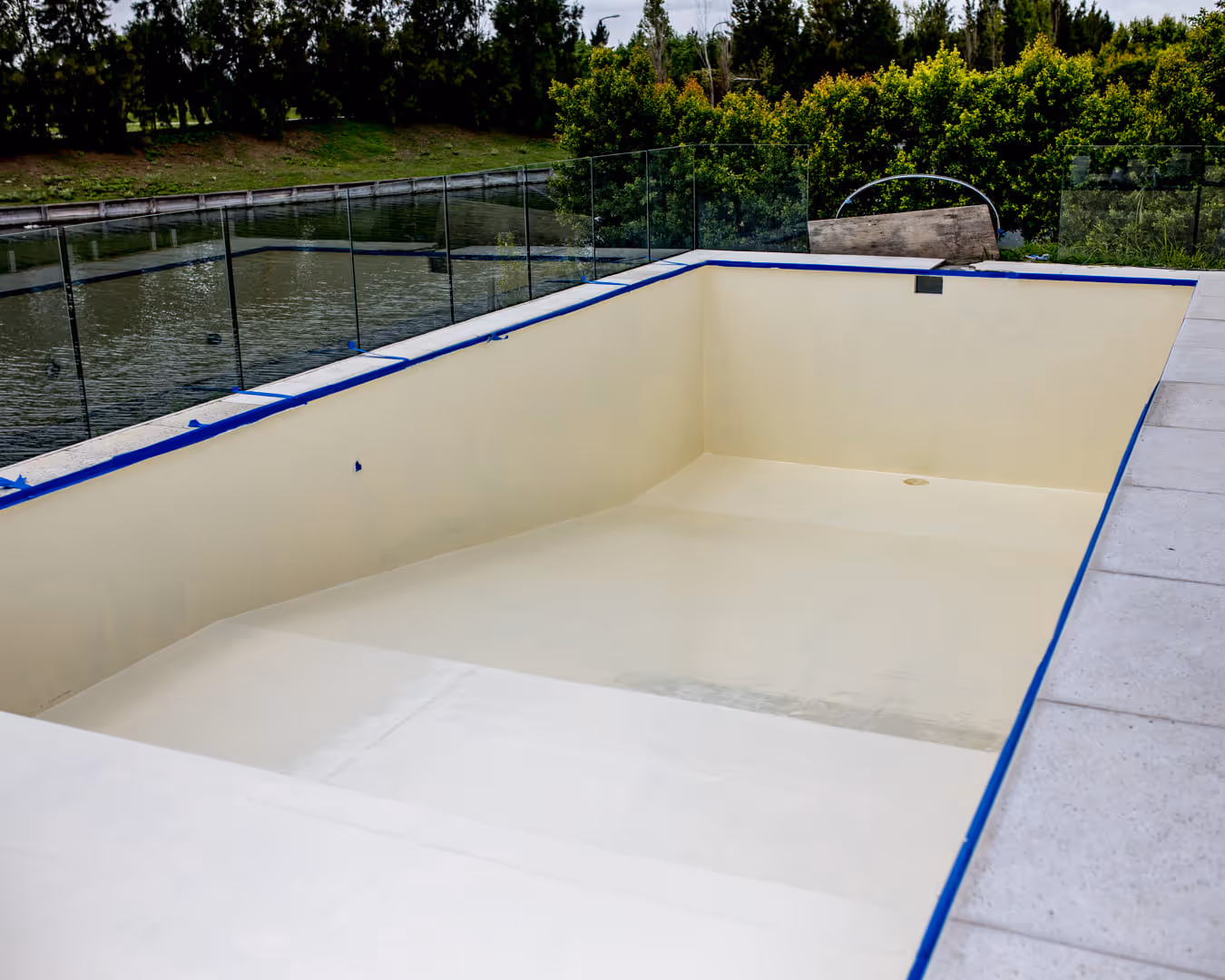 Piscina rectangular vacía con bordes de baldosas y valla de cristal, junto a un estanque y árboles.