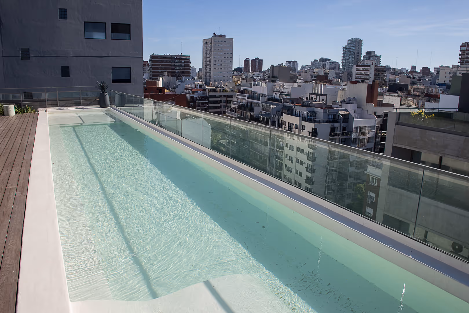 Moderna piscina en la azotea con agua cristalina y barandilla de cristal con vistas a los edificios de la ciudad bajo un cielo despejado.