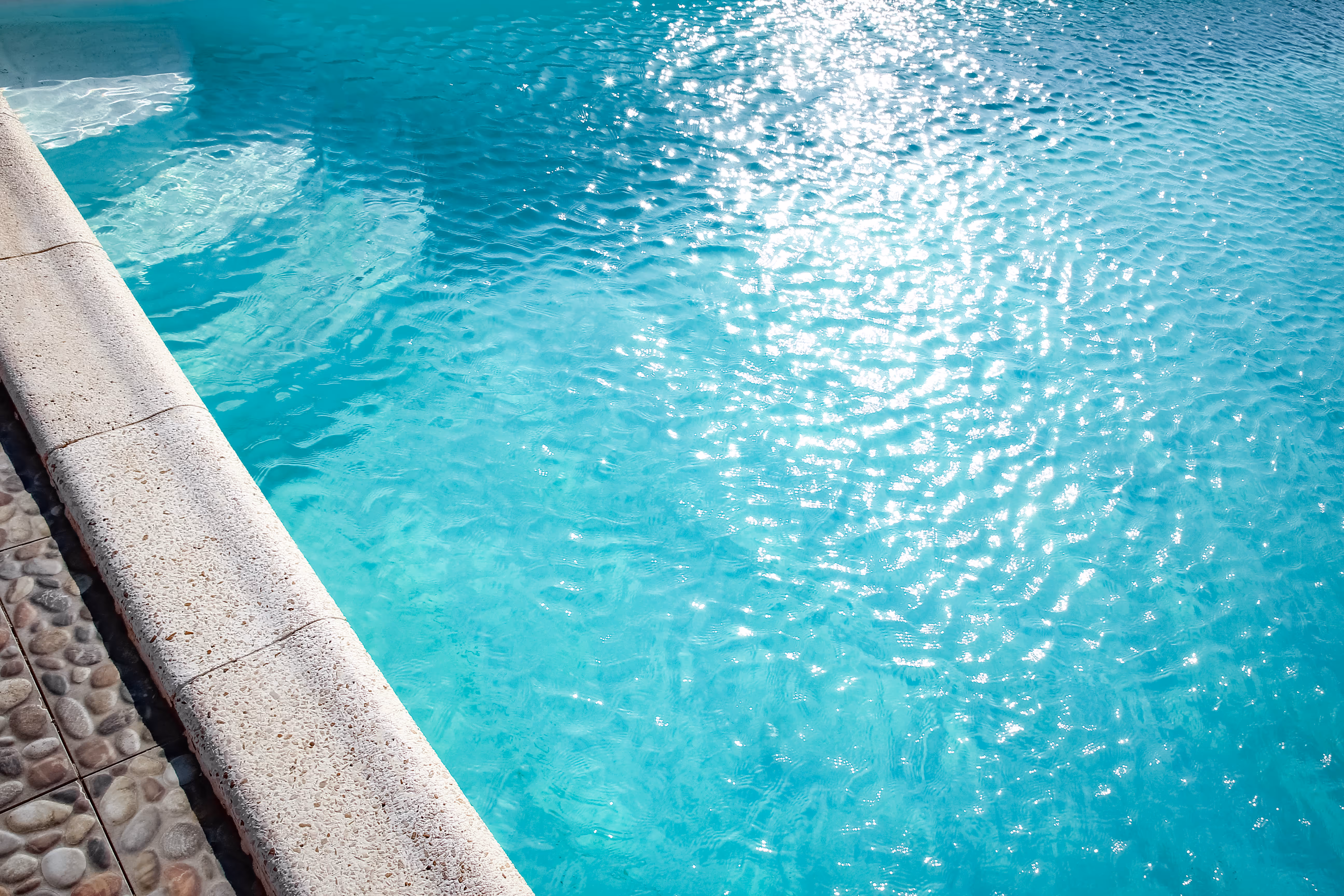 Primer plano de una piscina de agua turquesa clara con reflejos de luz solar y un borde de piedra.