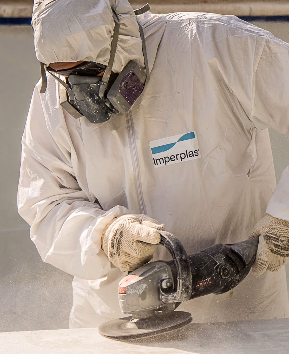Trabajador con traje de protección y máscara utilizando una amoladora angular de mano sobre una superficie.