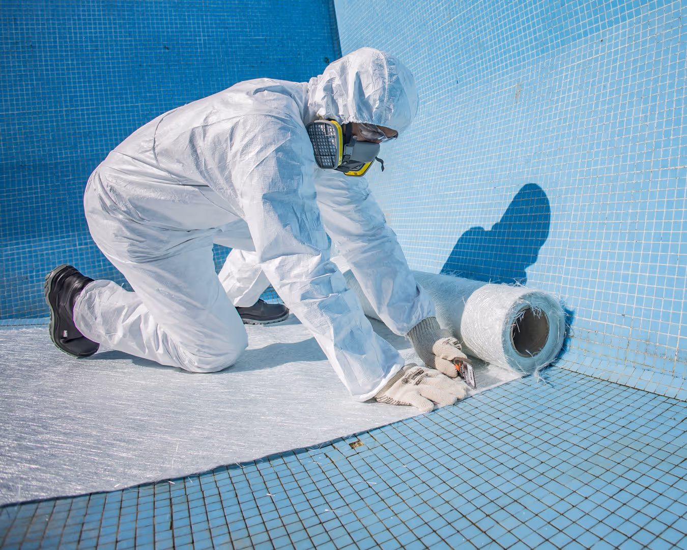 Worker in protective suit and mask laying fiberglass mat inside empty blue tiled pool for renovation.