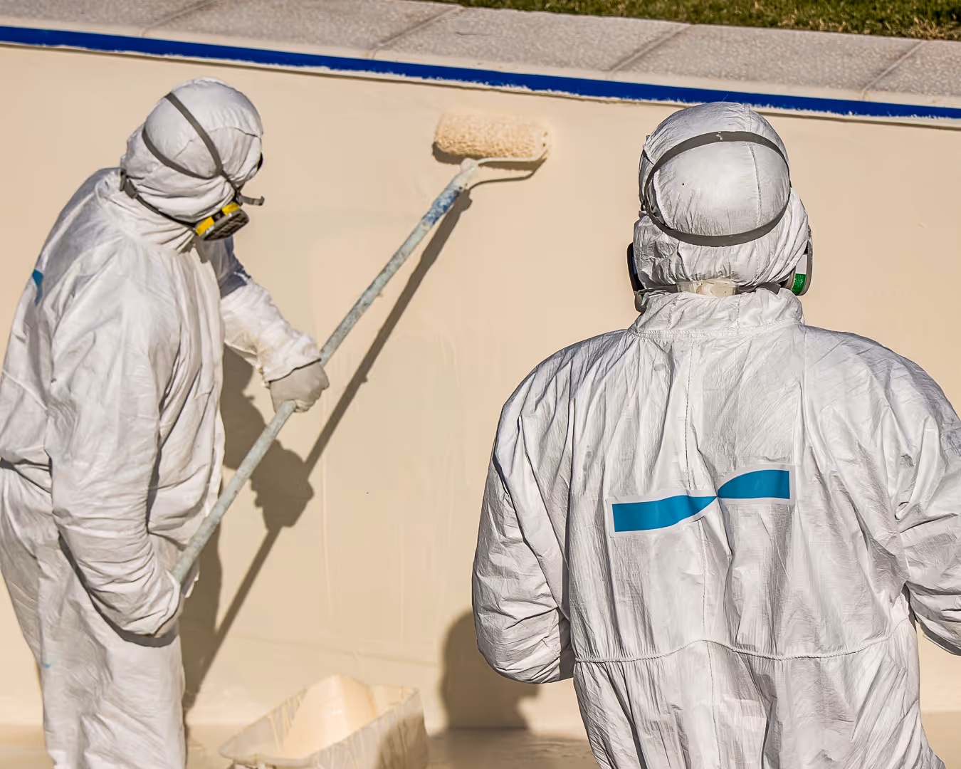 Two workers in protective white suits and respirators applying beige coating to a surface with a paint roller.