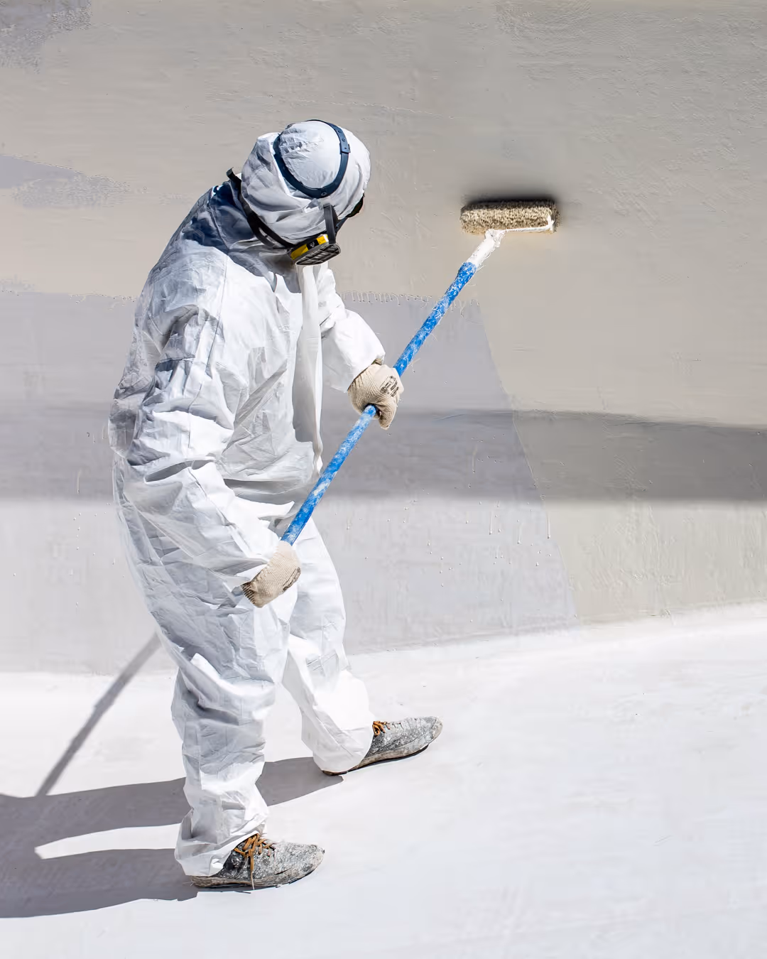 Person in full protective suit and respirator applying coating on a wall with a paint roller.