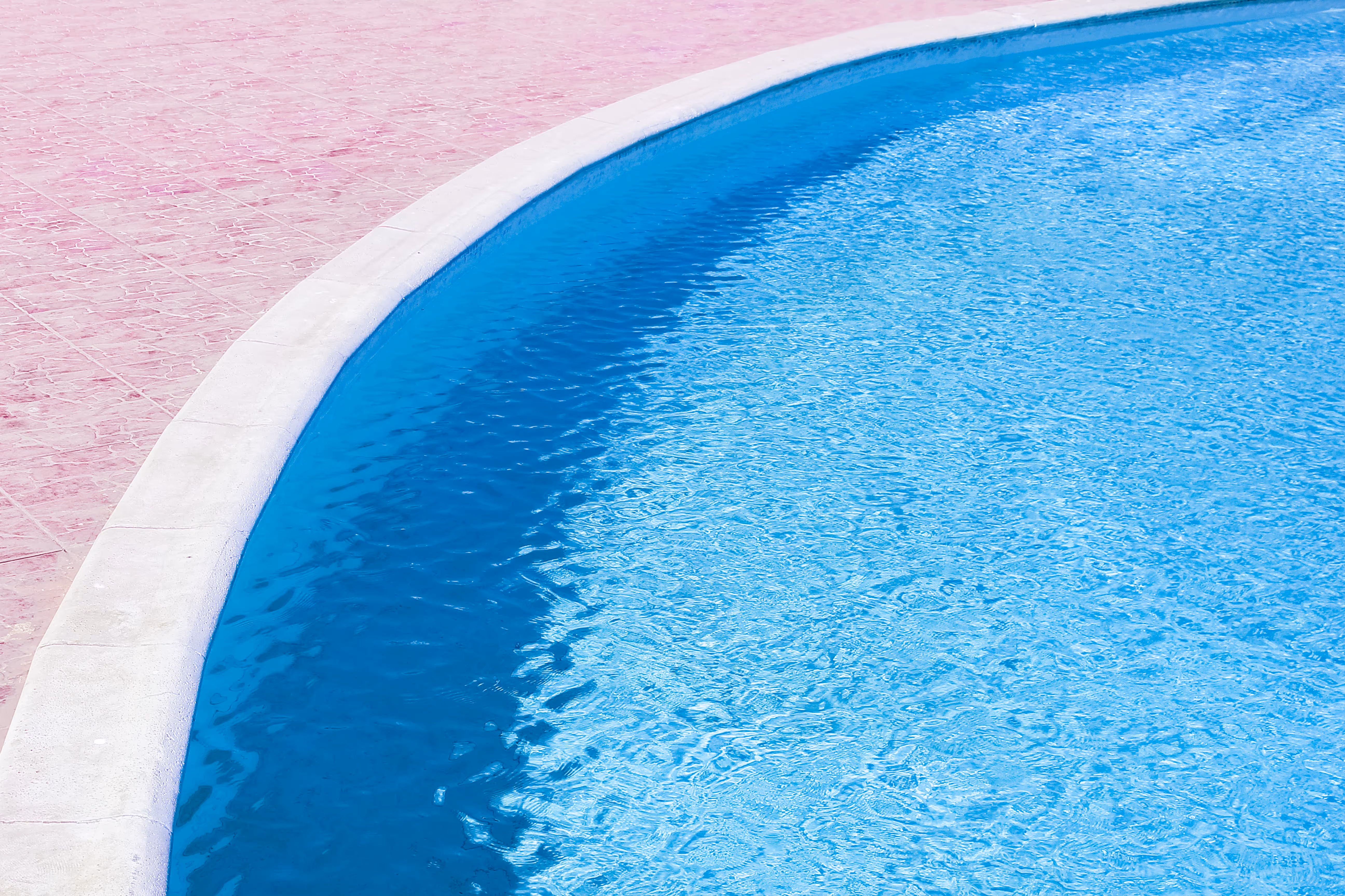 Curved edge of a blue swimming pool with a white border and pink tiled pool deck.