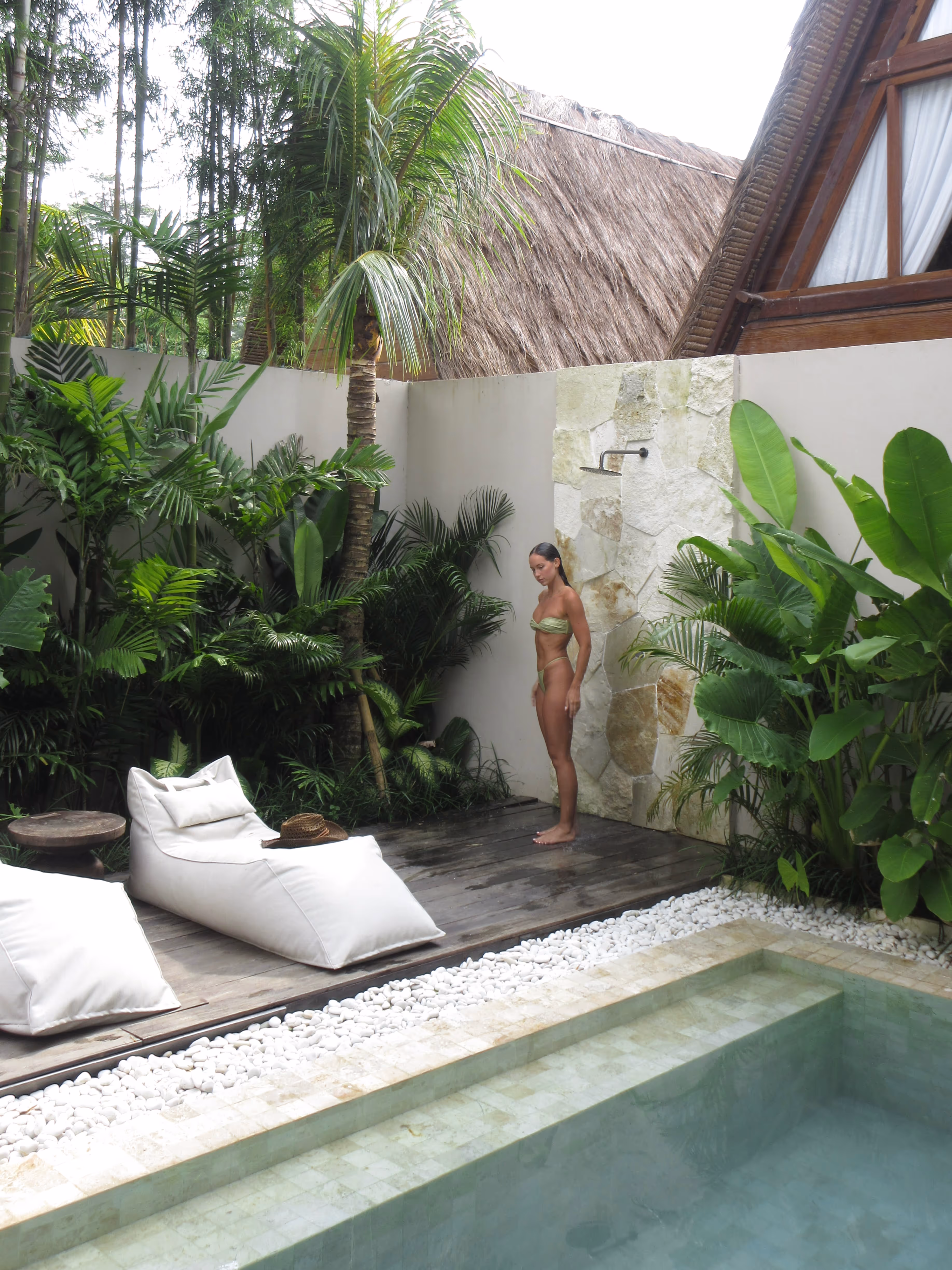 Woman in green bikini standing on wooden deck near outdoor shower surrounded by tropical plants and pool.