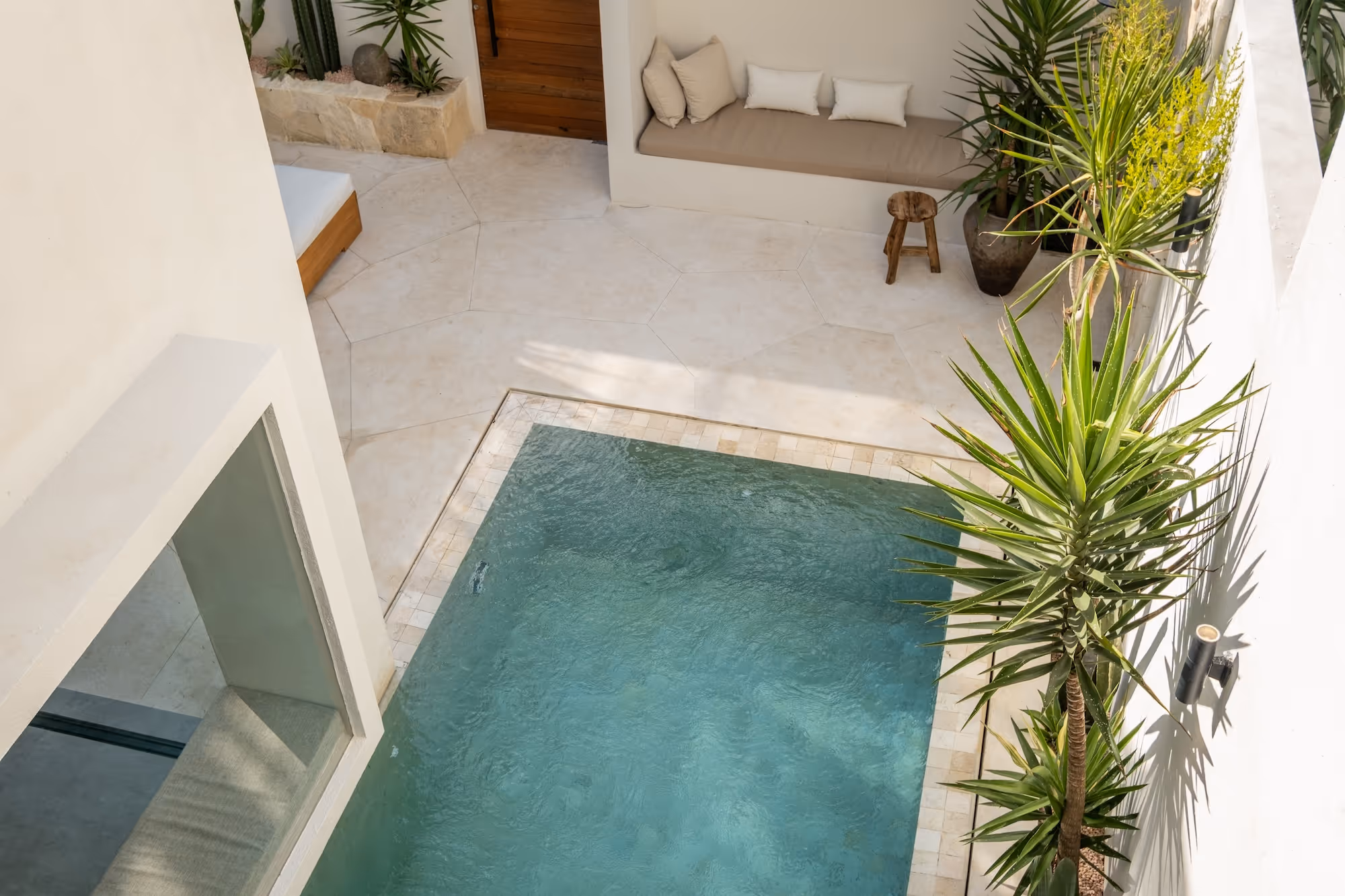 Top view of a small modern courtyard with a blue pool, white tiled floor, built-in seating with cushions, wooden stool, and tall green plants.