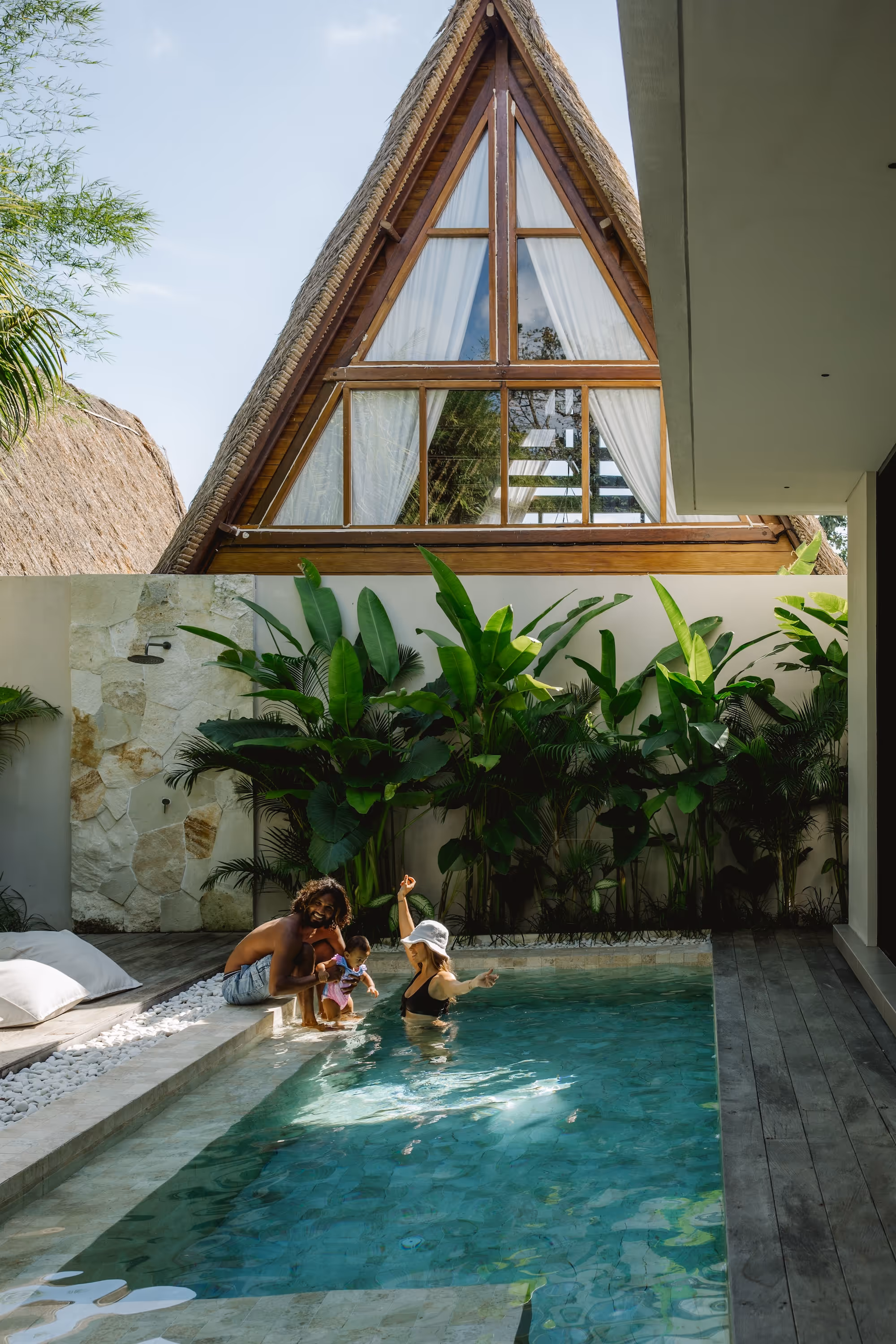 Family with a man, woman, and child enjoying time by a pool in front of a large triangular wooden house with lush green plants.