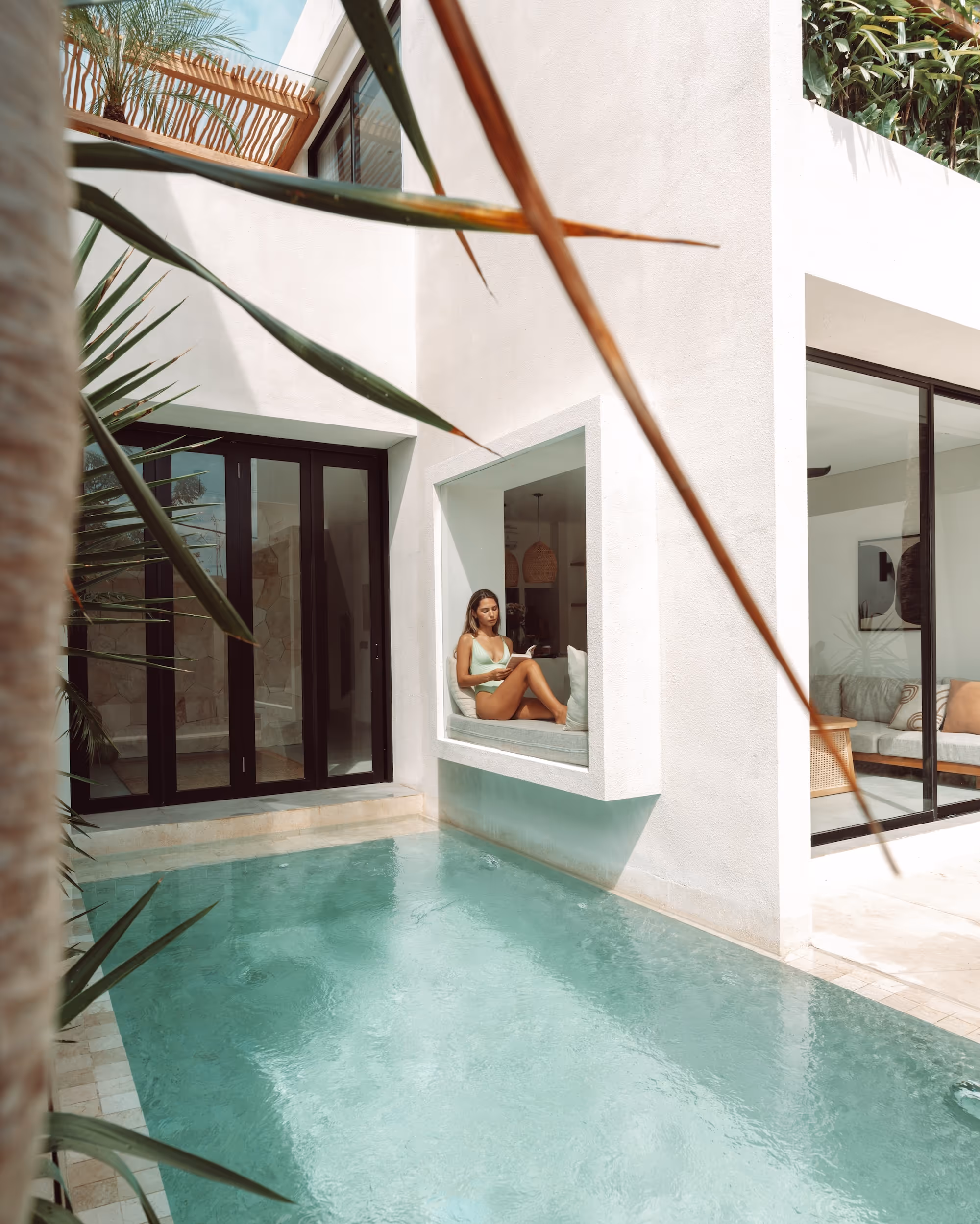 Woman in a light green swimsuit reading a book while sitting on a window seat overlooking a turquoise pool in a modern white house.