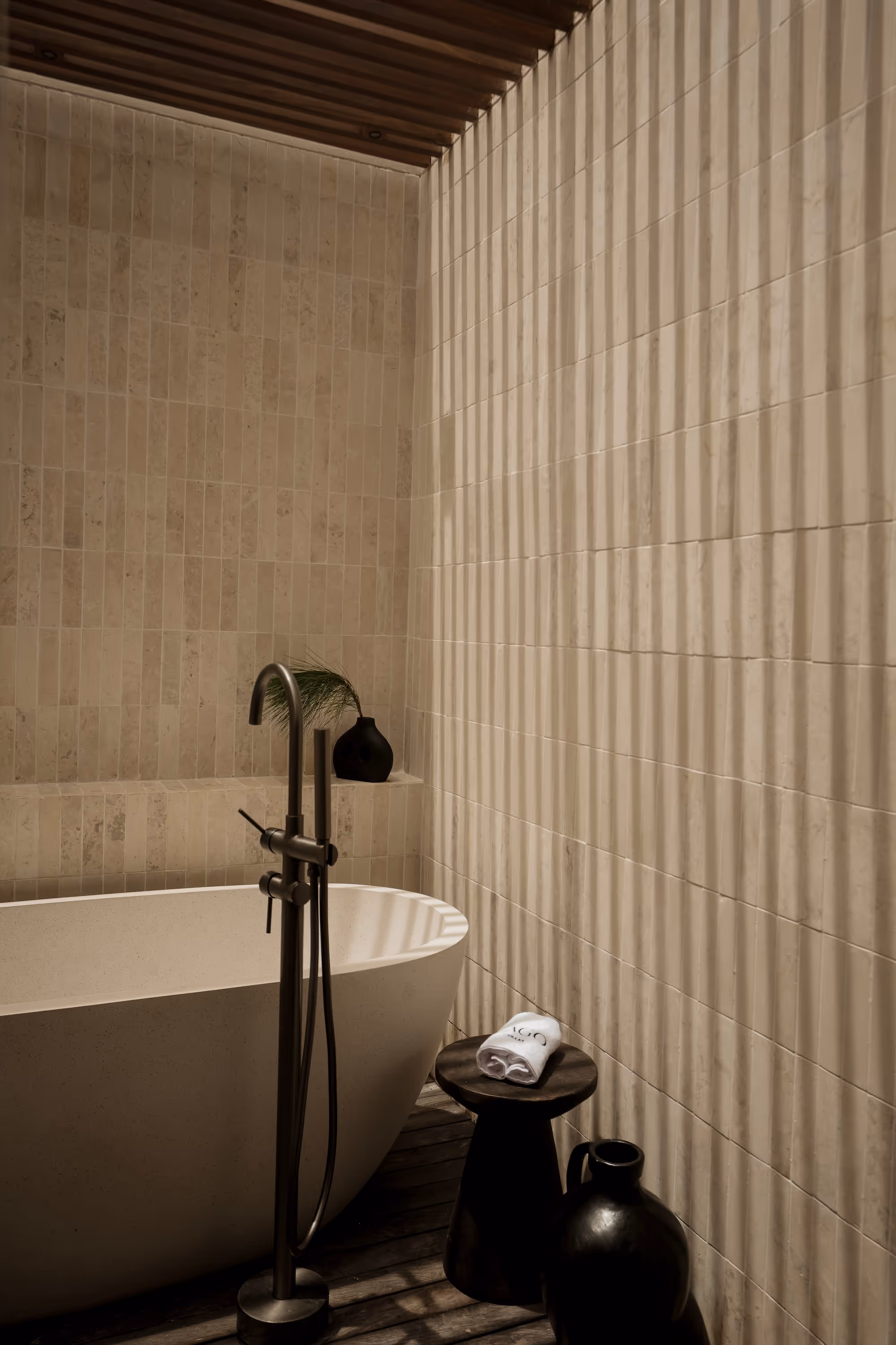 Minimalist bathroom with a freestanding white bathtub, dark floor-mounted faucet, wooden stool with a folded towel, and shadow stripes on beige tiled walls.