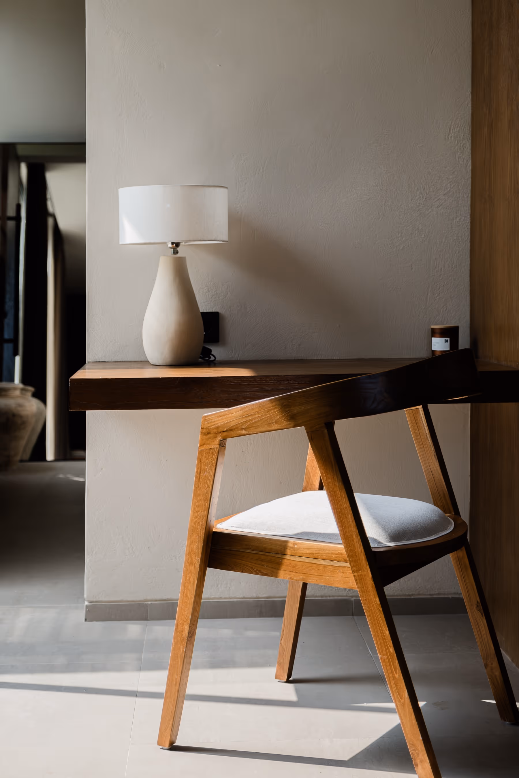 Wooden chair with white cushion next to a wall-mounted wooden desk holding a white table lamp.