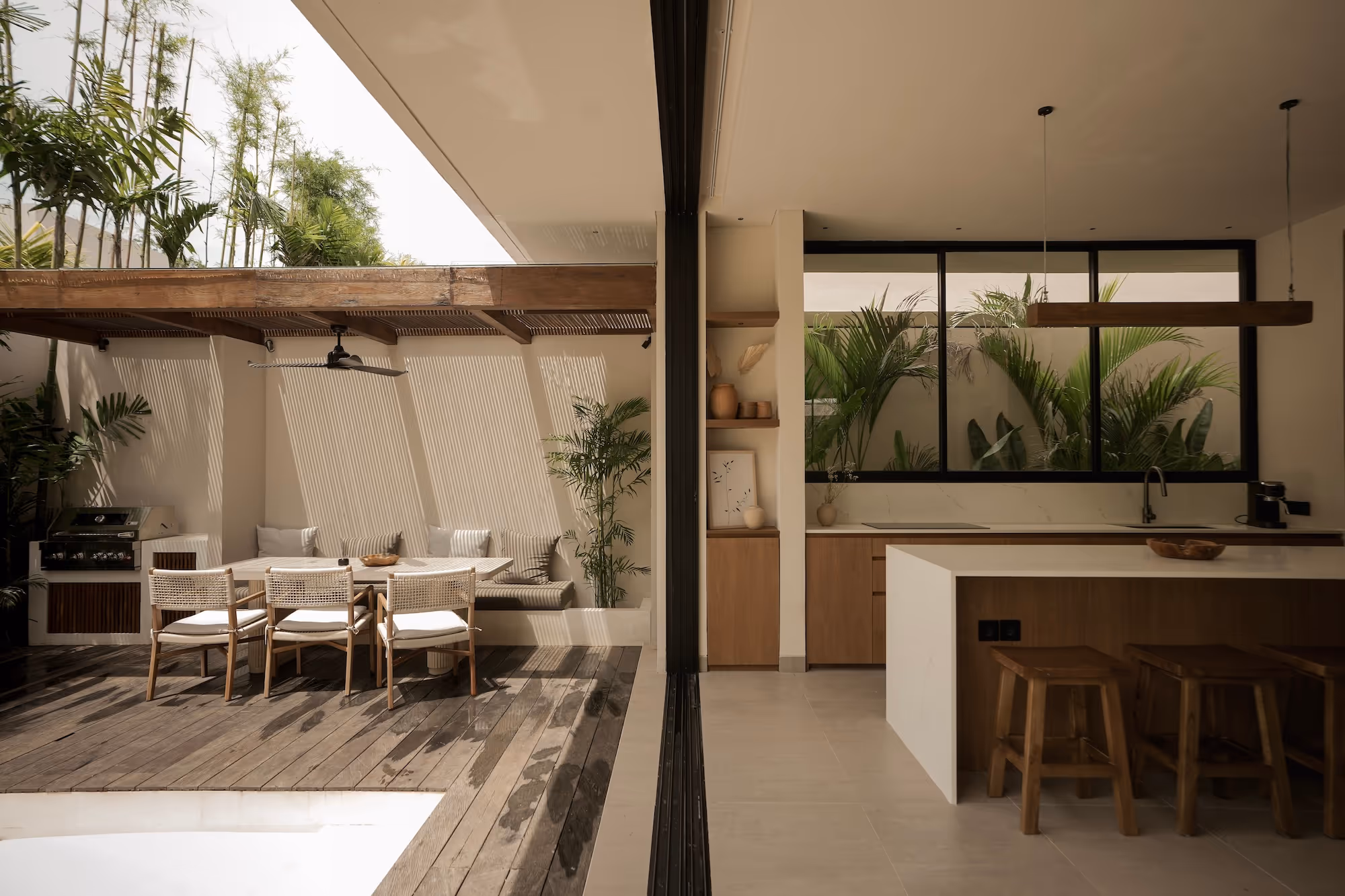 Modern open-concept outdoor dining area with wooden table and chairs next to a minimalist kitchen with wooden cabinets and indoor plants.