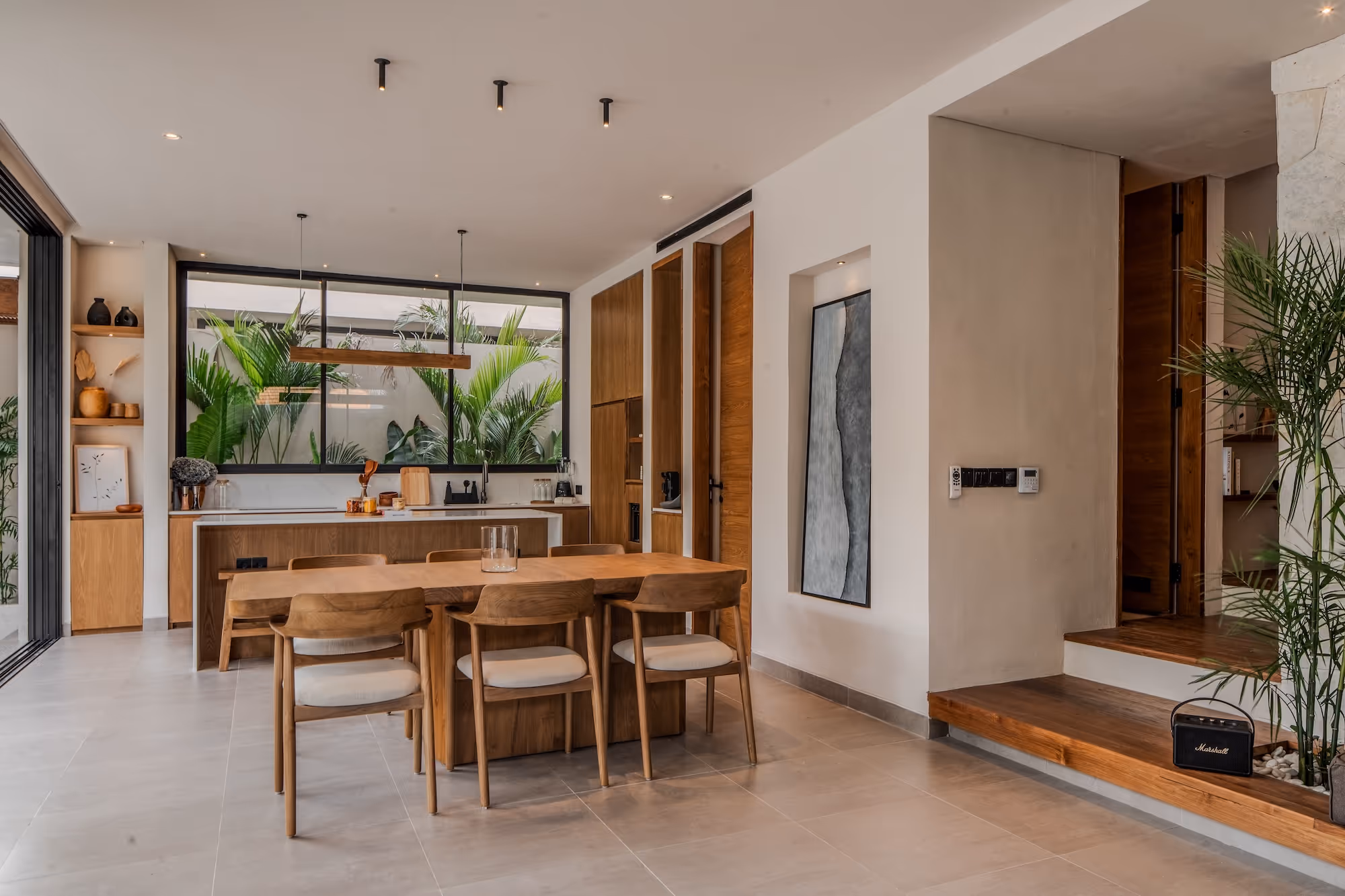 Modern dining area with wooden table, six chairs, large window showing lush green plants, and open kitchen with wooden cabinetry.