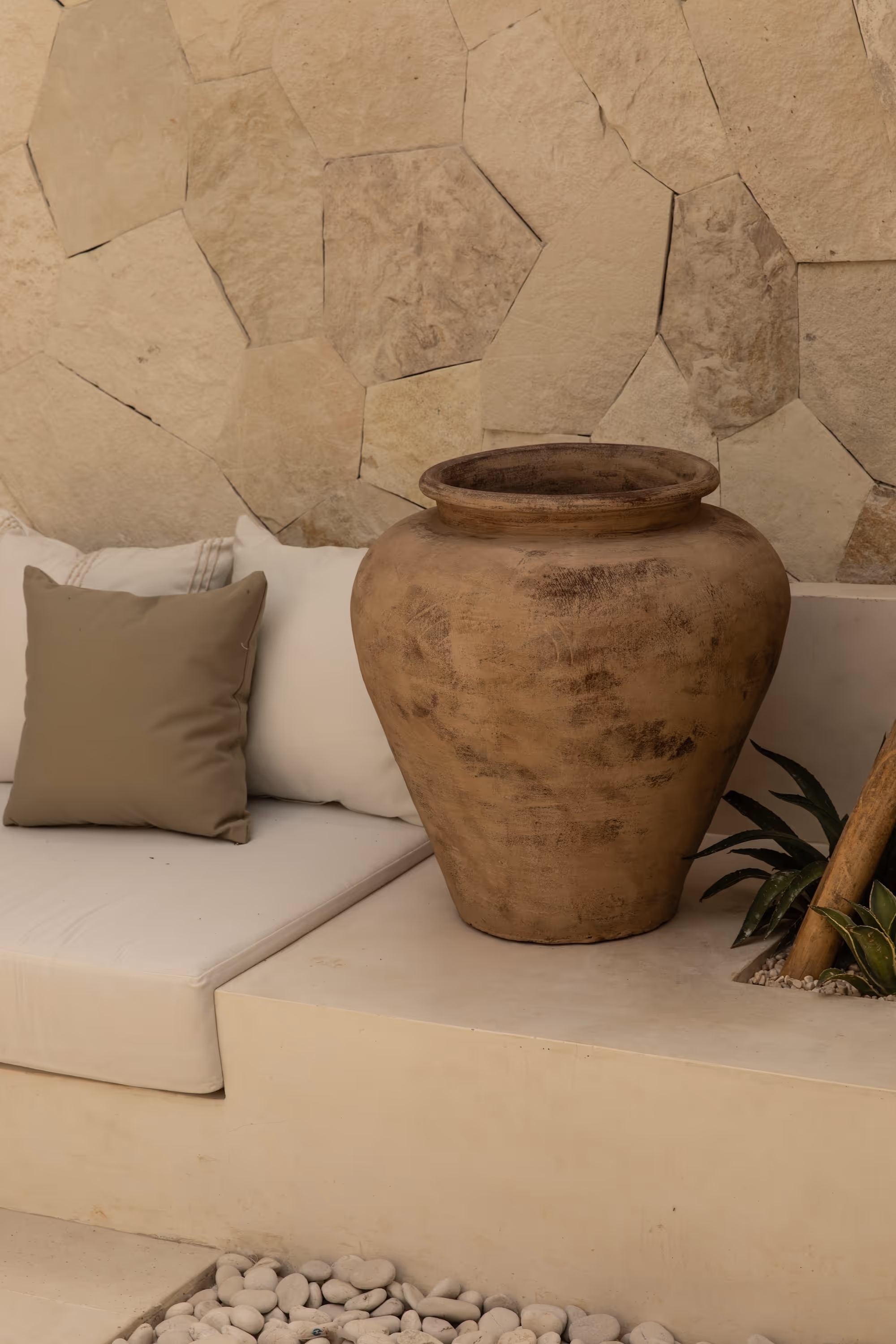Large rustic clay pot on a beige ledge next to a cushioned seating area with neutral pillows and a stone wall background.