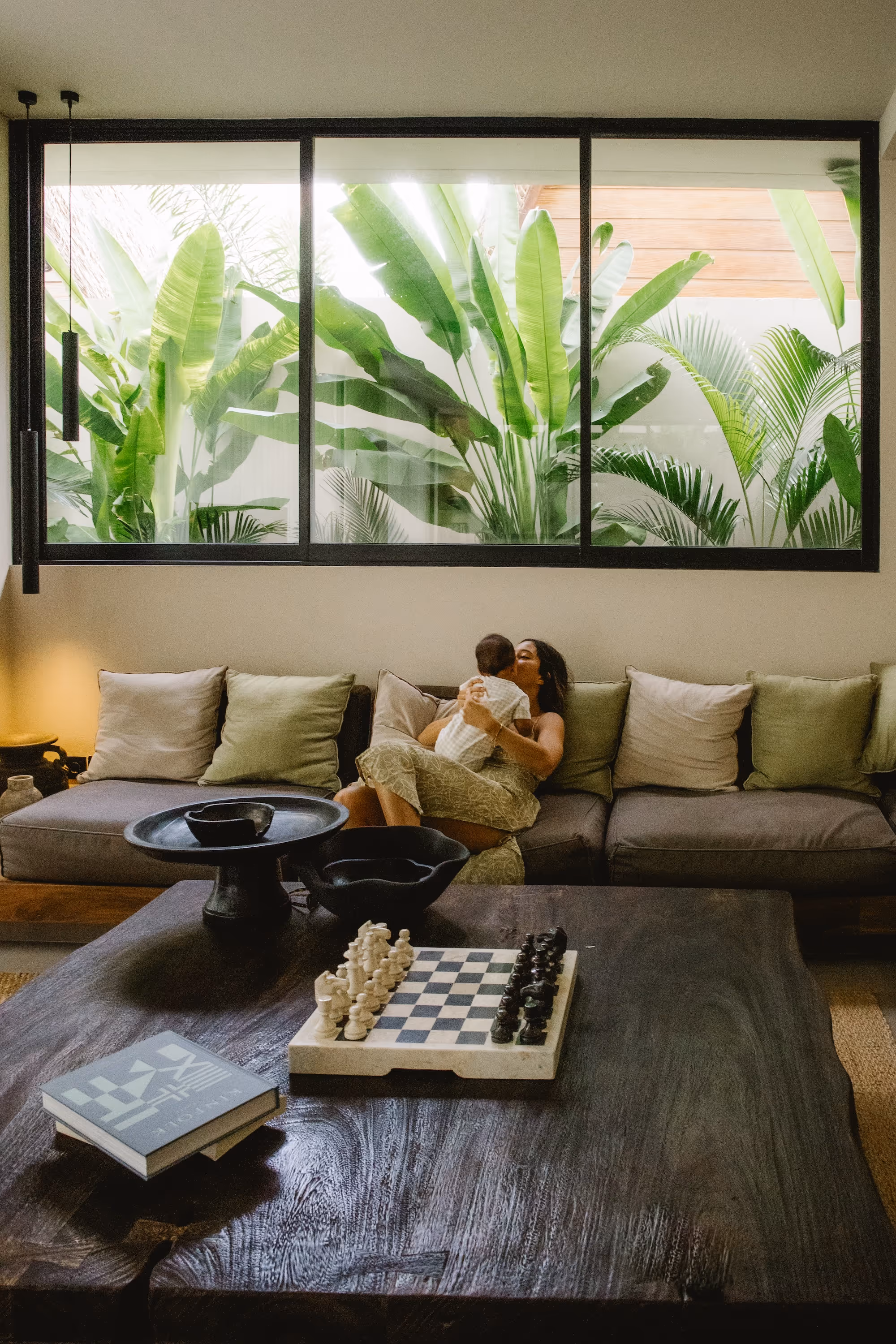 Woman sitting on a couch holding and kissing a baby in a cozy living room with large window showing lush green plants outside.