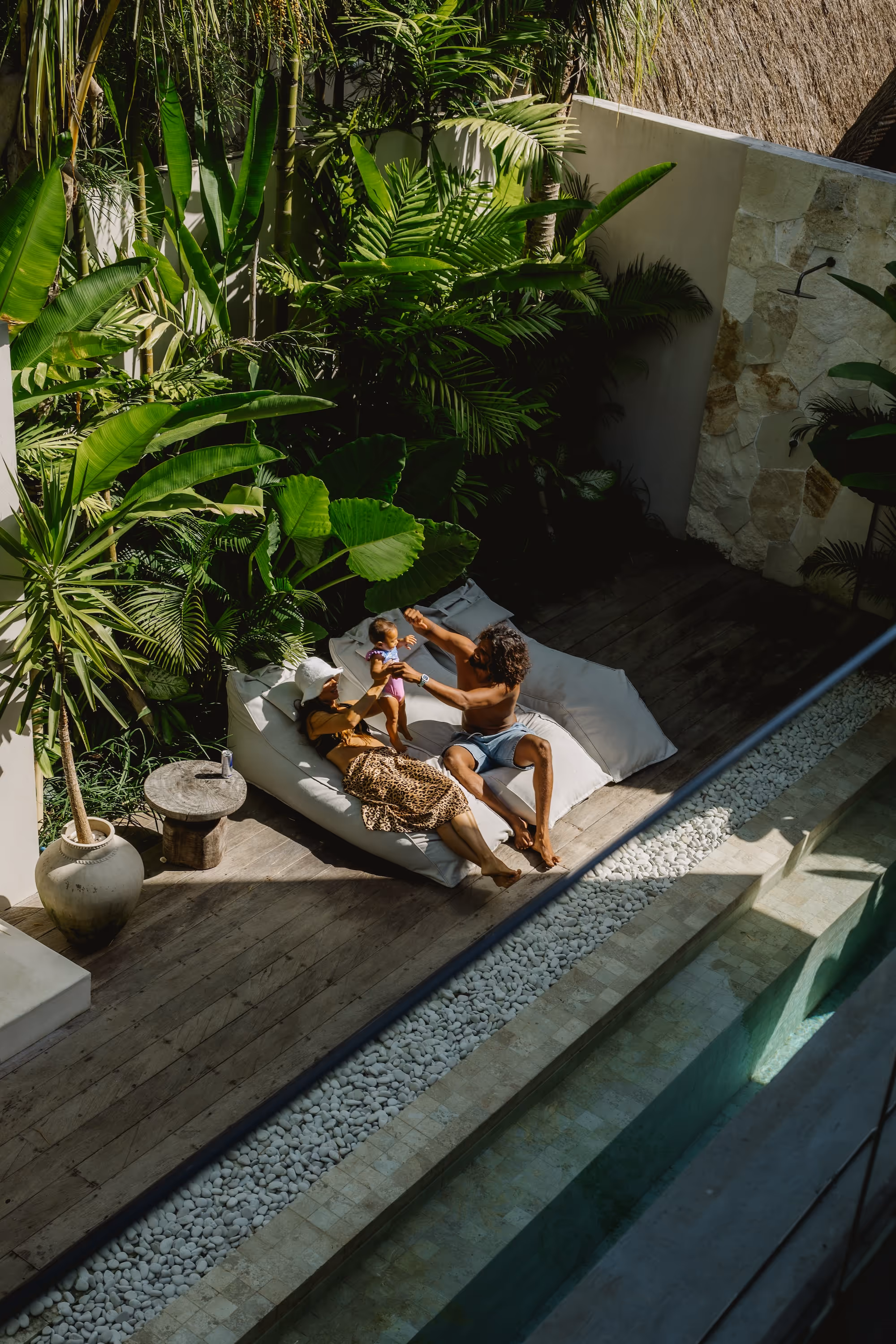 Family relaxing on outdoor white cushions near a pool surrounded by dense tropical plants.