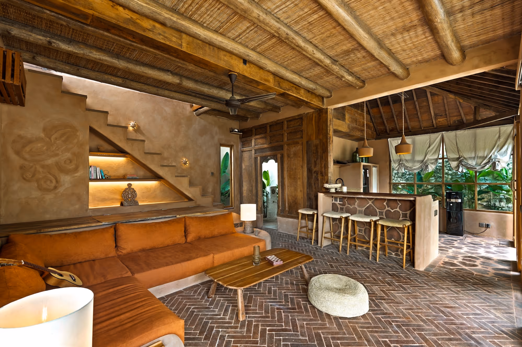 Rustic living room with a brown L-shaped sofa, wooden coffee table, woven pouf, bar stools, exposed wooden beams, and large window with hanging curtains.