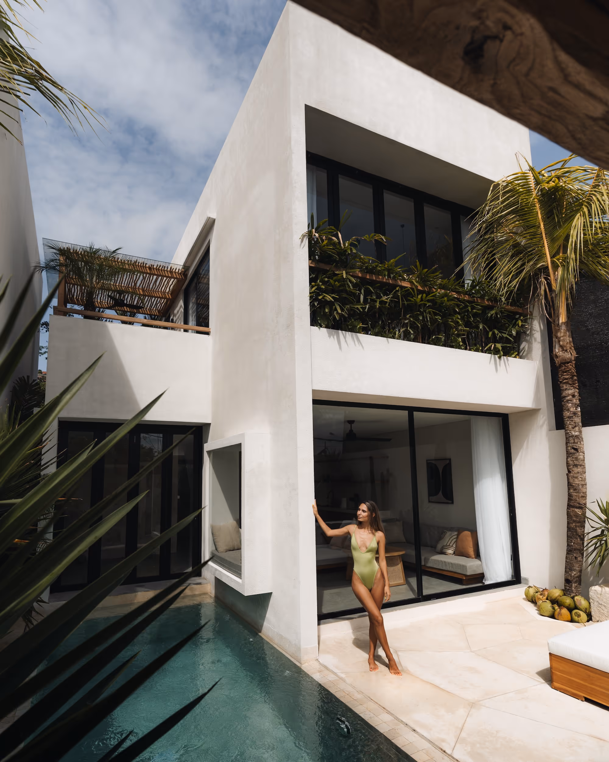 Woman in a green swimsuit standing by a modern white house with a pool and palm trees.