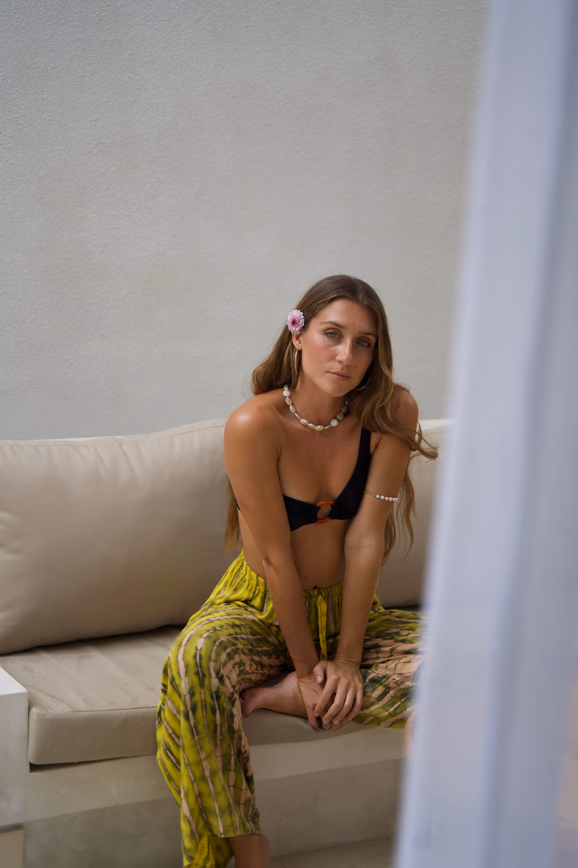 Woman with long hair wearing a black bikini top and yellow patterned pants sitting on a beige couch with a pink flower in her hair.
