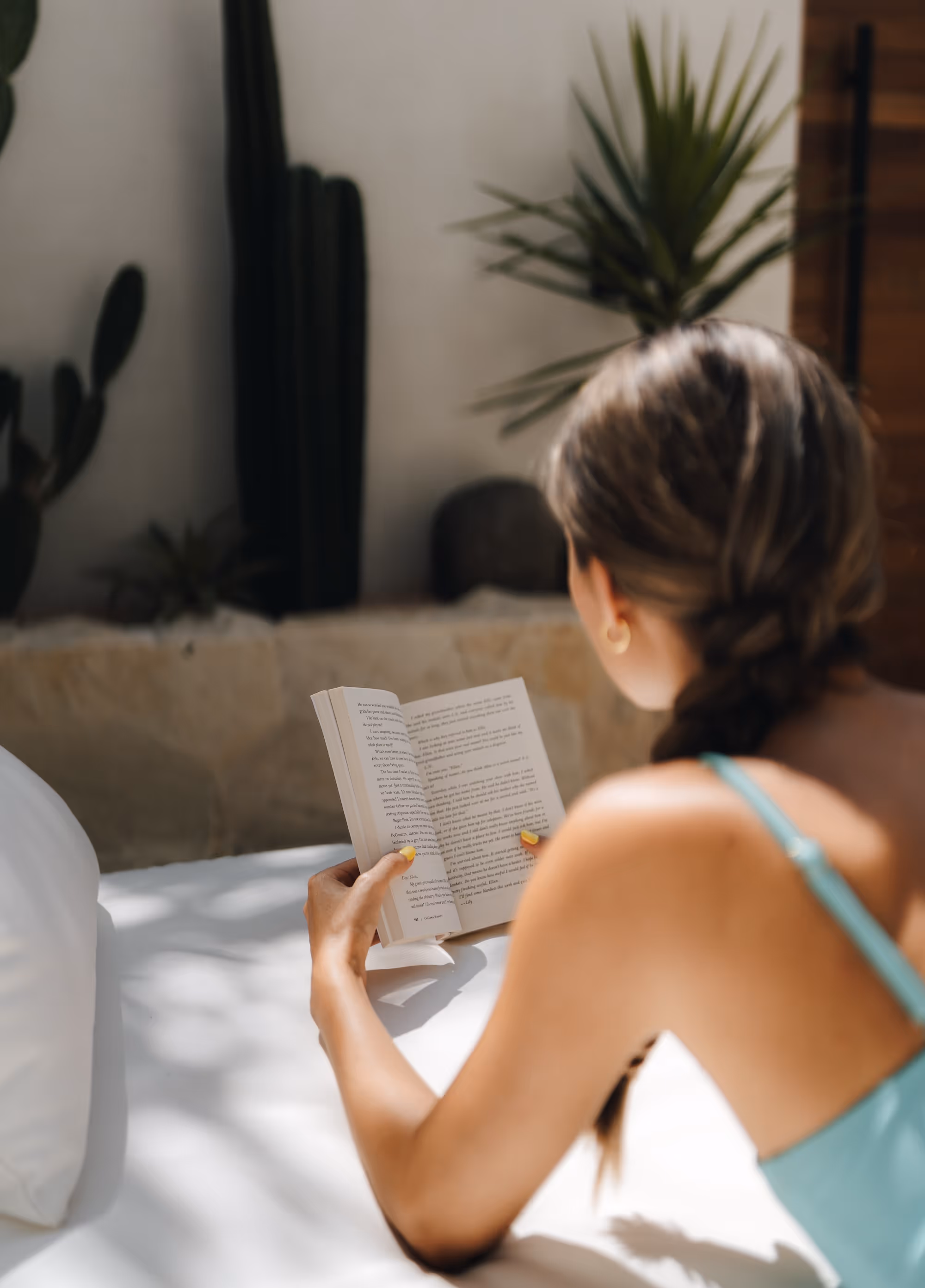 Woman with braided hair in a teal top reading a book while lying on a white bed in a sunlit room with plants in the background.