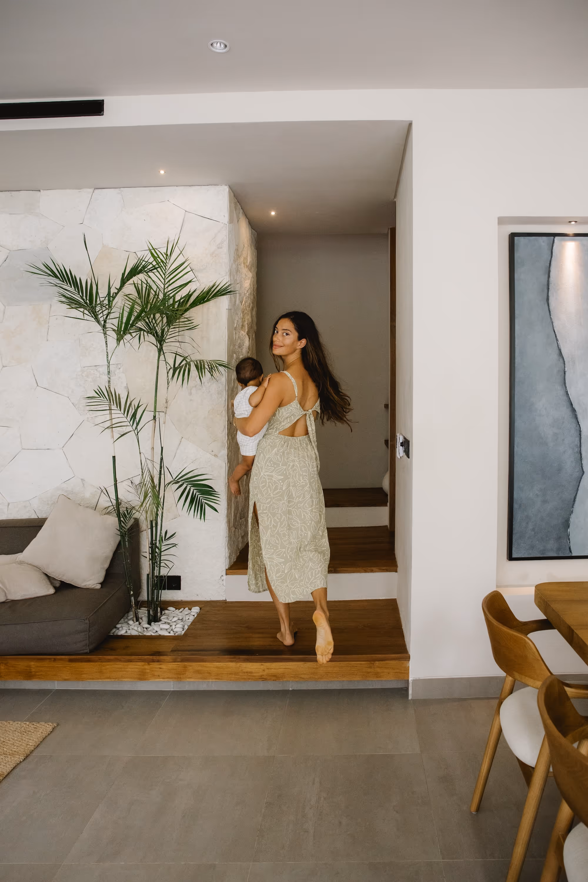 Woman in a light green dress holding a baby while walking barefoot up wooden steps inside a modern home.