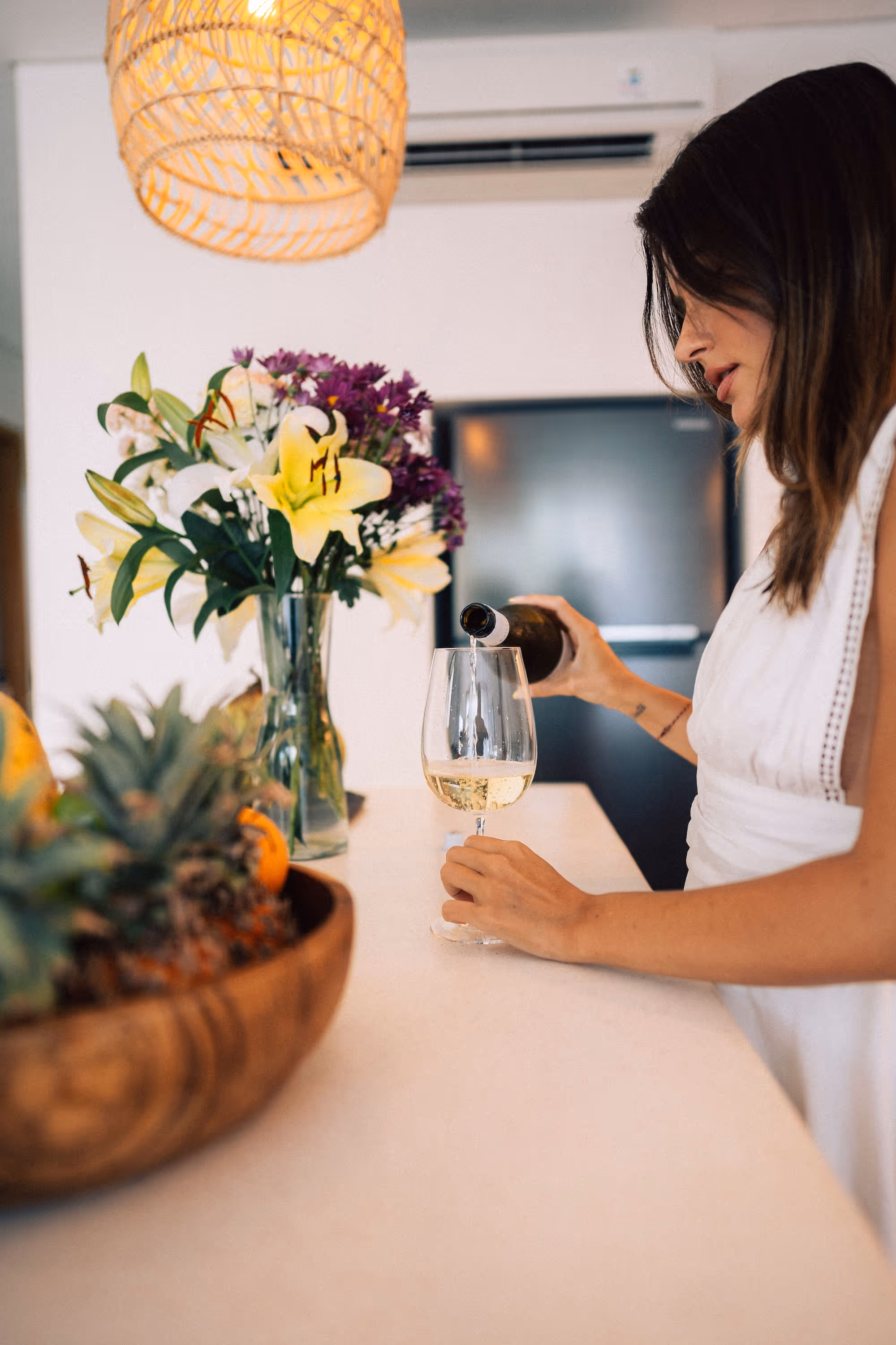 Woman in white dress pouring white wine into a glass at a countertop with a vase of flowers and a bowl of fruit.
