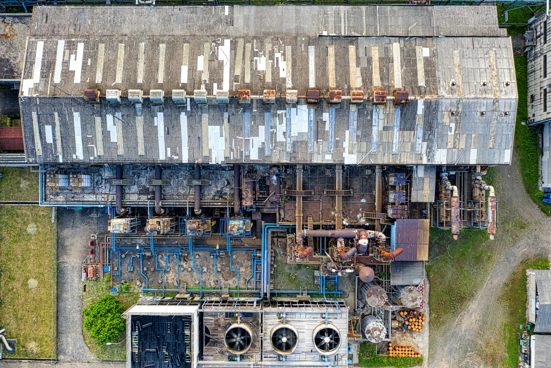 Aerial view of an industrial facility showing a large building with a weathered roof, connected piping, and machinery.