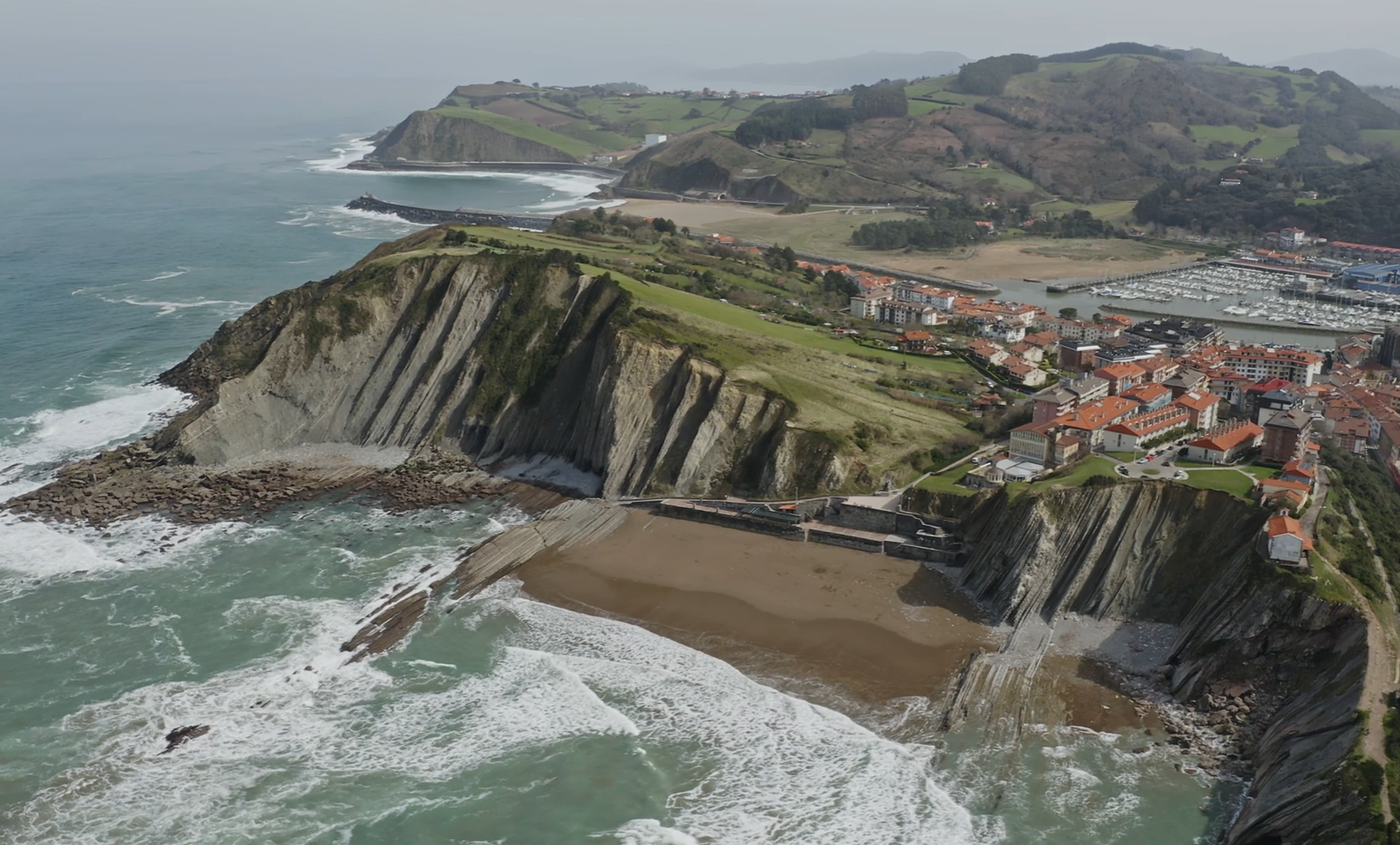 Aerial view of cliffs and a sandy beach with waves, adjacent to a coastal town with red-roofed buildings and a marina.