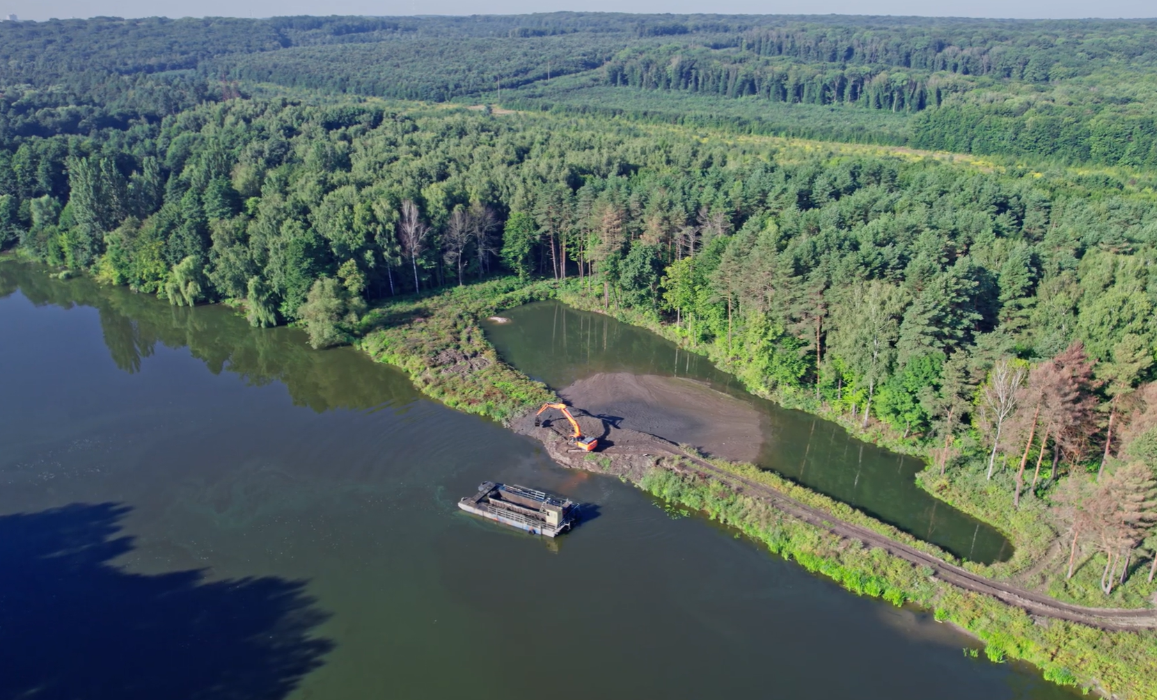 Aerial view of a lake beside dense forest with an excavator dredging near the shore and a floating barge in the water.
