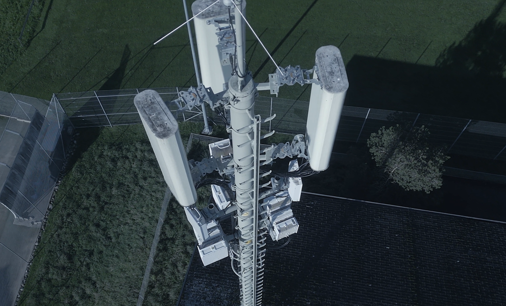 Aerial view of a cellular tower with antennas and equipment mounted on it, surrounded by grass and fencing.