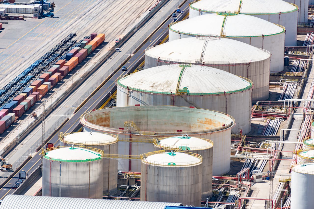 Aerial view of large circular industrial storage tanks and a railway with cargo containers beside a road.