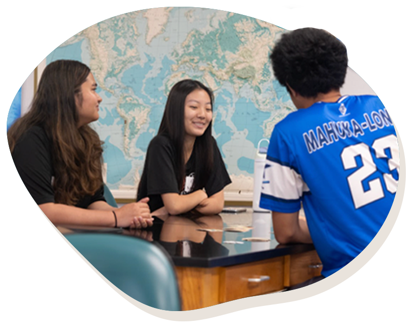 Three students sitting at a table in a classroom with a world map on the wall behind them.