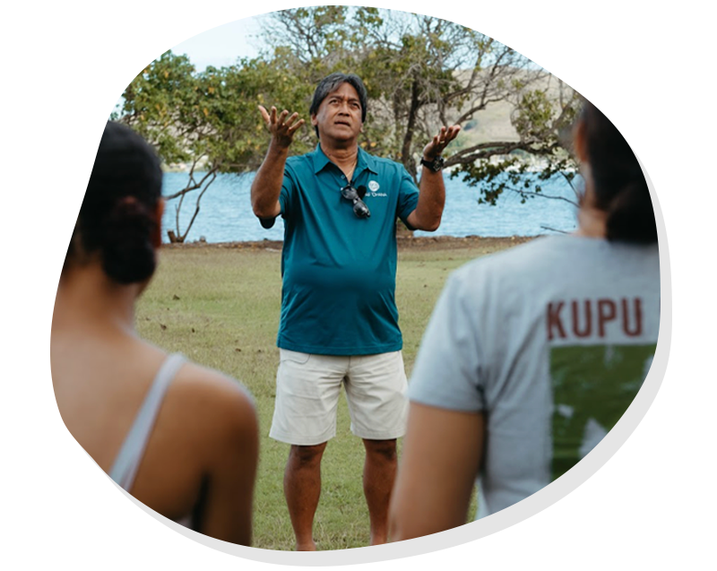 Man in blue shirt speaking outdoors to two people near water with trees in the background.