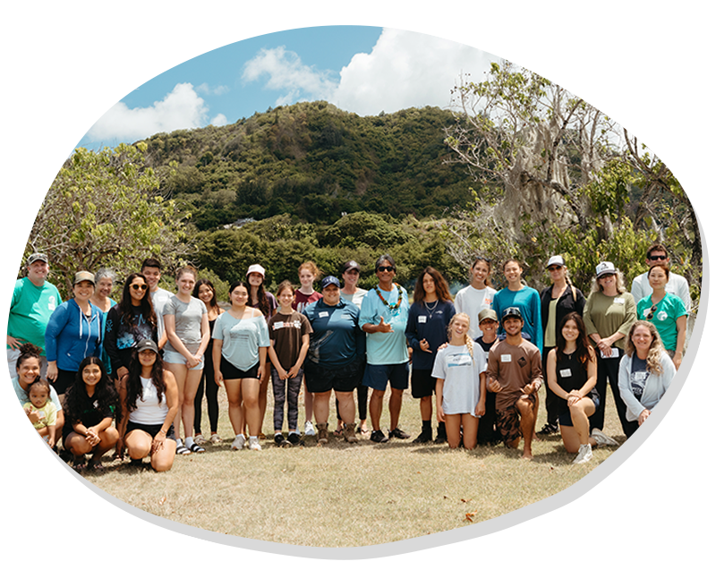Diverse group of people posing outdoors with trees and a forested mountain under blue sky.