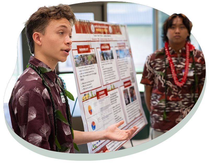 Young man in patterned shirt presenting research poster while another listens in similar attire with lei.
