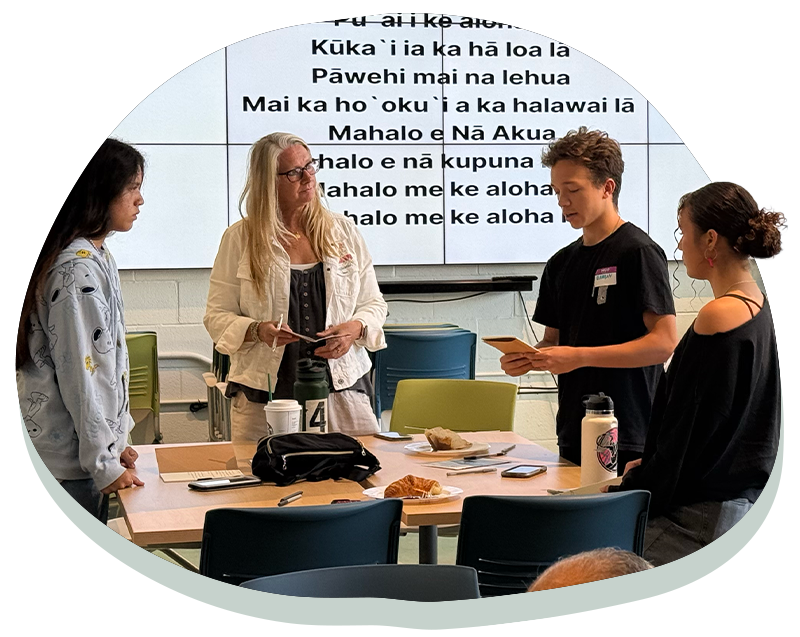 Four people standing around a table during a discussion, with text displayed on a screen behind them.