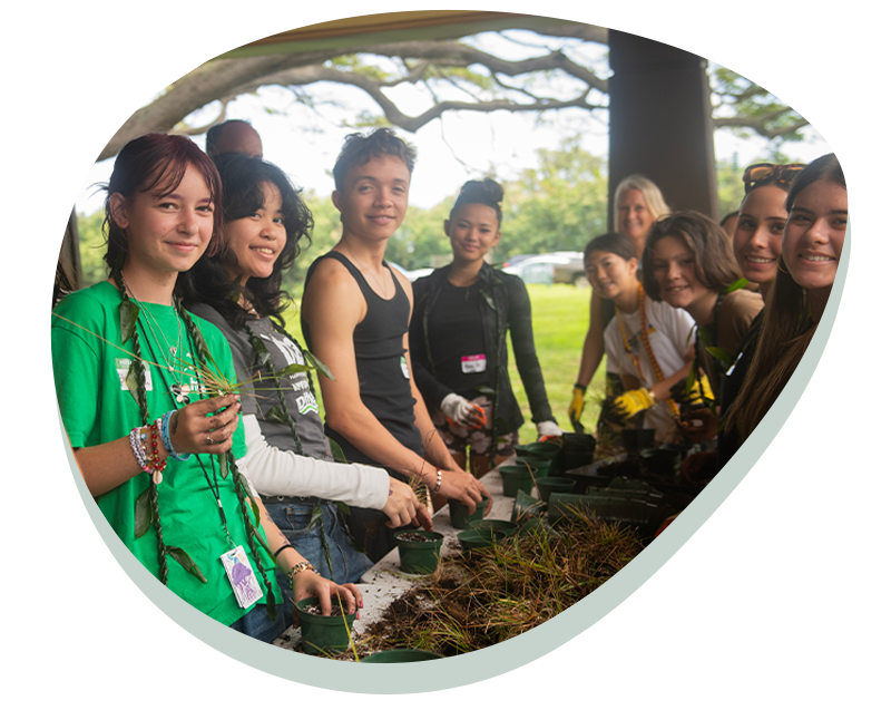 Group of young people planting seedlings in small pots at an outdoor table under a tree.