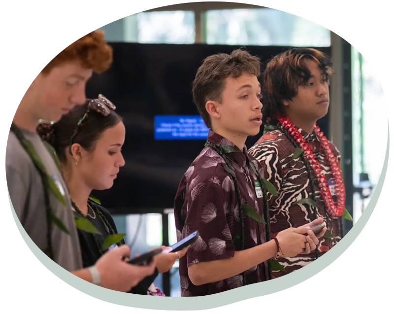 Four young people standing in a row, three holding phones, one speaking, all wearing leis indoors.