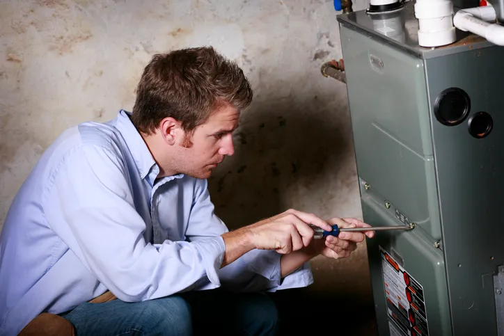 A professional HVAC technician in a light blue shirt and jeans is kneeling and using a screwdriver to open a residential furnace.
